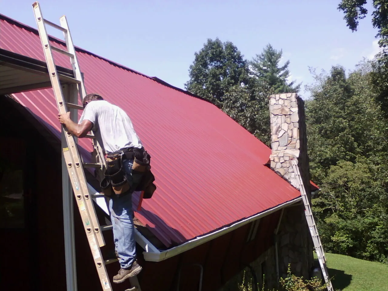 Roofer on a ladder beside a red metal roof and stone chimney — Soffit & Fascia Repair by Roofing Brothers in Coquille